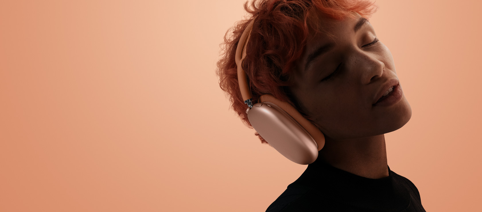 オレンジのAirPods Max 2を装着している人物。オレンジがかったピンク色の背景を背に、頭を傾けて目を閉じている