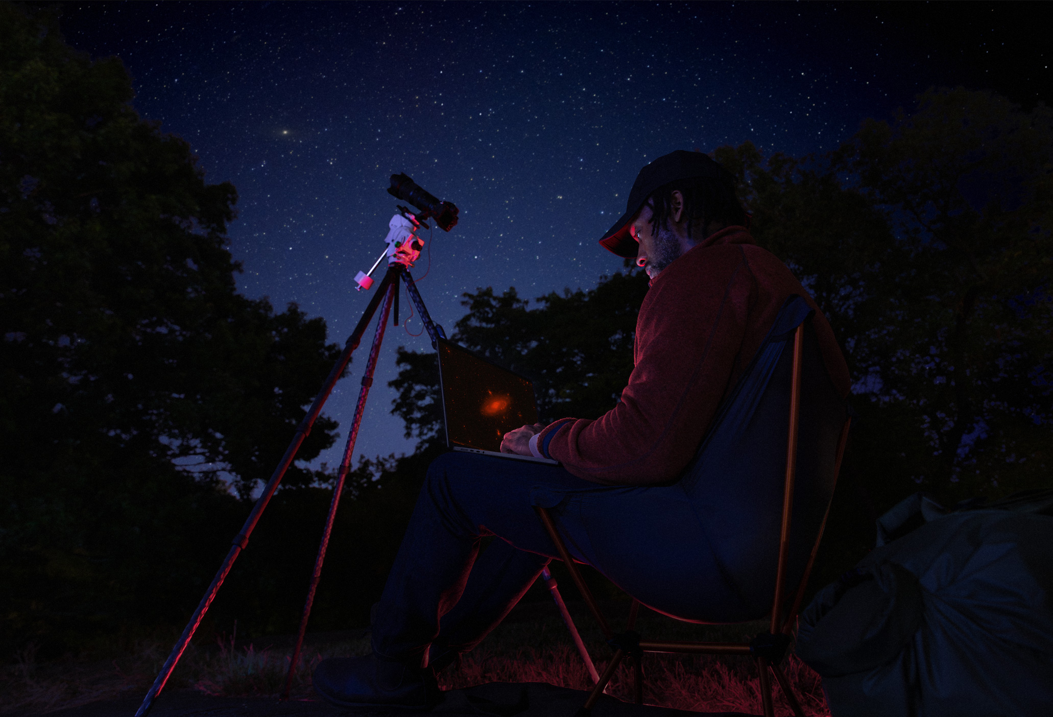 夜に屋外でMacBook Proを膝の上に置いて使っている人物。MacBook Proは星空に向けられた三脚の上のデバイスに接続されている